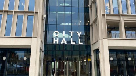 Sunderland City Hall. It is a tall, modern building, made of beige panels and a lot of glass. There is metal City Hall writing above the revolving doors at the entrance. 