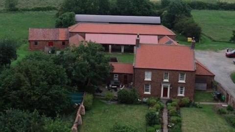 An aerial view of a rural property with a red‑roofed brick house, several outbuildings and a landscaped garden surrounded by fields.