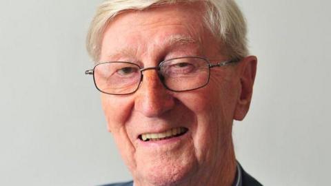The picture shows an older man sitting against a plain light background. He has short, neatly combed white hair and is wearing thin, rectangular glasses. He is smiling in a warm, relaxed way. 