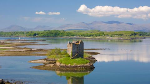 Seann chaisteal cloiche, le tùr, agus pòla brataich, air eilean beaga ann an loch-mhara. Tha eilean nas motha le craobhan air am meadhan an deilbh agus beanntann air cùl sin fada às. Chithear faileas dhen chaisteal agus na sgòthan san uisge.
