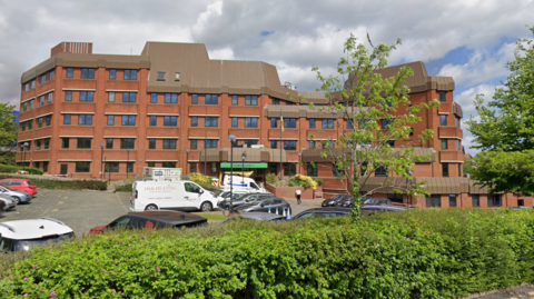 A multi-storey red brick buildings with a car park in front of it. At the entrance a green sign reads "Town Hall".