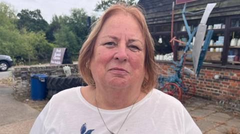 A woman with shoulder-length ginger hair. She is wearing a white t-shirt and standing next to what looks like a barn.