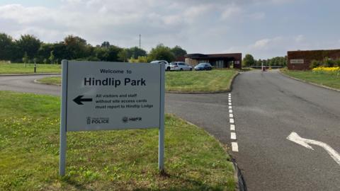 A white sign with two legs is positioned on a stretch of grass near a private road. The white sign reads: "Welcome to Hindlip Park."