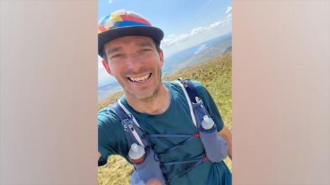 Selfie of Pete Faulkner. He is smiling into the camera as he stands on a hill. He is wearing a multi-coloured baseball cap and a blue T-shirt.