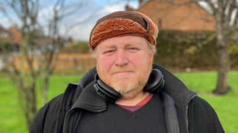 A bearded man in a trapper hat looks sombrely into the camera. 