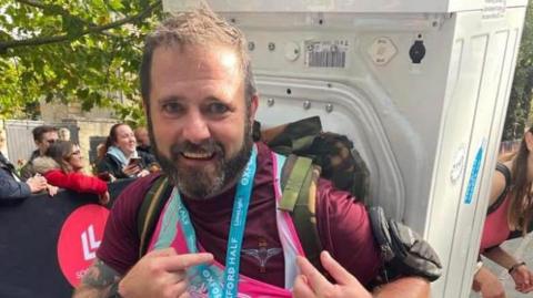 Martin has short brown hair and a brown beard. He is at the end of his half marathon, with a washing machine strapped to his back. He is pointing to a medal around his neck. 