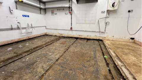 A kitchen has been ripped out of a room that has had the floors removed due to flooding. The flooring is brown. Kitchen appliances and shelves remain on the far wall. 