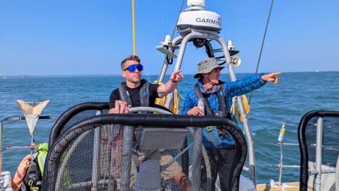 Kevin Lynch is standing behind the sailing wheel, he has one hand on the wheel and the other pointing to the front. He is wearing a black t-shirt and sunglasses.