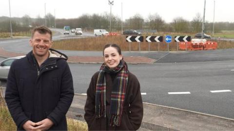 A man with short, light brown hair, wearing a blue parka coat stands next to a woman with a dark brown ponytail wearing a brown coat and tartan scarf in front of a roundabout.