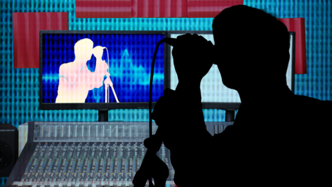 A black shadow of a male is singing into a microphone in a music studio. A music desk sits below a screen projecting the shadow onto a computer screen. 