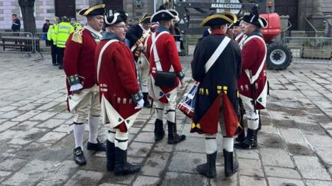 Members of Jersey Militia at a previous year's commemoration event. The men are dressed in uniforms which are red and white. They are standing on a paved road. Man in high-viz jackets are standing near railings in front of buildings in the background.