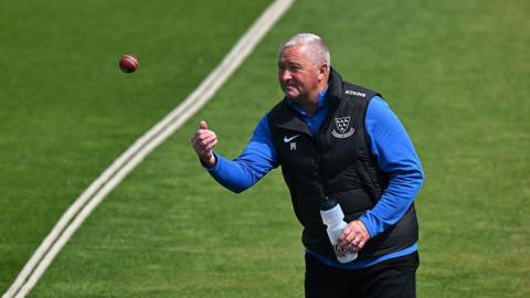 Paul Farbrace throws a ball underarm near a boundary rope in training with a water bottle in his other hand