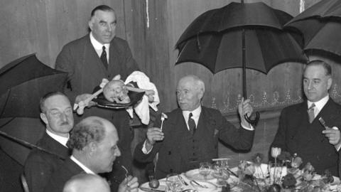 Archive photo from 1934 showing men sitting at an elaborately-decorated dining table while holding open umbrellas above their heads