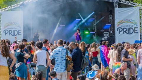A performer on a stage in a park. A large group is watching them. There are two white banners either stage of the stage with the words "CAMBRIDGE PRIDE" on them.