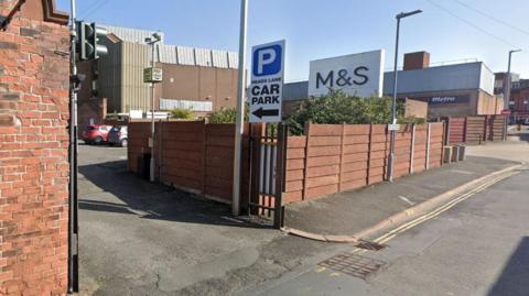 Google Maps image of the entrance to Heads Lane car park. A white sign has a large blue logo with a white letter "P", with Heads Lane Car Park written underneath. An M&S shop can be seen in the background along with a Tesco Metro.