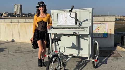 Chloe Marie Aston in a yellow cardigan and black dress stands alongside her piano bike.