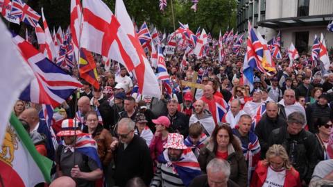 Dozens of people are seen at the Unite the Kingdom march in central London. Many England and Union Jack flags can be seen in the foreground and background. Some of the men and women in the crowds can be seen wearing Union Jack flags around their shoulders