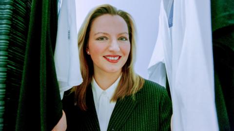 Michelle Mone in pin-striped business jacket and white shirt in front of a rack of white shirts. She is smiling broadly and has long blonde hair.