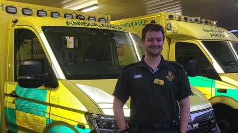 The image shows a male paramedic standing in front of two ambulances.