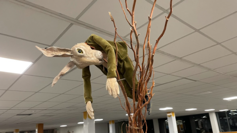 A rabbit puppet wearing a green velvet suit hanging from a branch inside a large room.