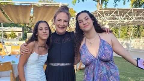 Yahel, left, wearing a white dress with mother Lianne, wearing black in the centre, and sister Noiya in a purple summer dress. They are all smiling while stood outside with a garden with tables and chairs in the background.