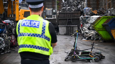 A police officer in high viz uniform stands in front of a pile of e-scooters at a scrapyard