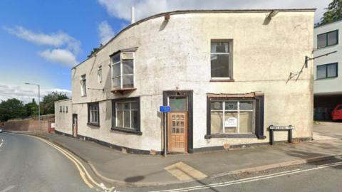 A dilapidated white-painted building stands on a street corner in Worcester. It has a curved front wall and a wooden door. A pavement runs around it and there are double yellow lines. The window is covered and there is a sign fixed to the glass.