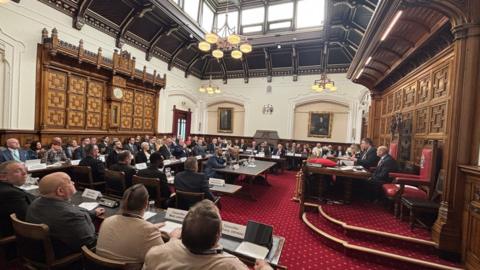 Council chamber with high ceilings, a red and gold carpet and seats filled with politicians wearing suits and smart business attire.