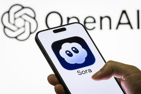 A person holding a phone with the Sora logo. It is a cloud with eyes visible on the screen against a white backdrop with the OpenAI logo displayed.