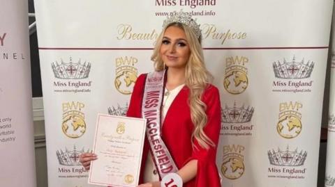 A woman wearing a white dress with a red jacket is smiling holding a certificate, wearing a Miss Macclesfield sash. She is standing in-front of a sign saying Miss England that is covered in crowns. 