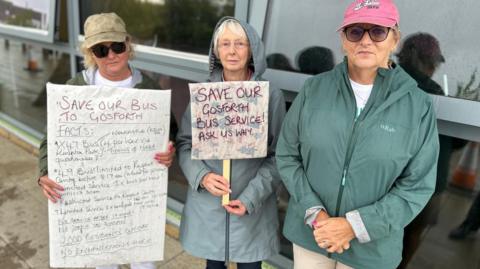 Three older women, with two holding signs opposing the axing of a bus service. One says 'Save our Gosforth Bus Service! Ask us why...'