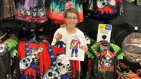 Etienne, wearing a white T-shirt holding his costume which is red and blue with a skull hanging from the front