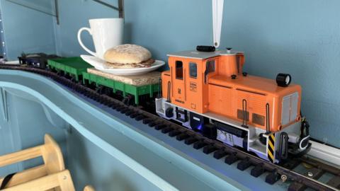 A small orange model train engine on tracks indoors pulling a bacon sandwich and a mug