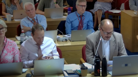 A full council meeting, with members sitting at rows of light wood desks with laptops and microphones in front of them. Five councillors' faces, four men and a woman, are visible, with more men and women partly visible behind them. 