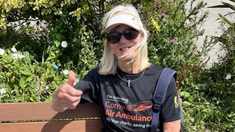 A woman wearing sunglasses, a cap and a 'Cornwall Air Ambulance' t-shirt holds a thumbs up to the camera while sitting on a bench in the sun.
