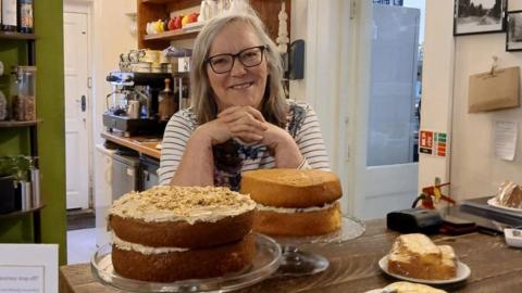 Lizzie Leigh smiles at the camera, wearing glasses and stripy top. She stands behind a wooden coffee bar stacked with cakes