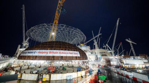 A view of Hinkley Point C being built. There are cranes and scaffolding arond the building. A banner on the building says: "Hinkley Point C - Helping Britain Achieve Net Zero"