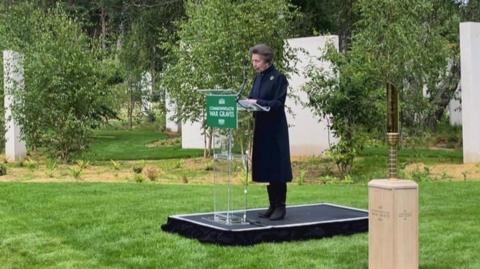A woman in a dark  coat and boots stands on a podium surrounded by trees and large upright stone slabs.
