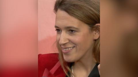 A woman, Dr Filby, has long brown hair and wears a black top with a red jacket