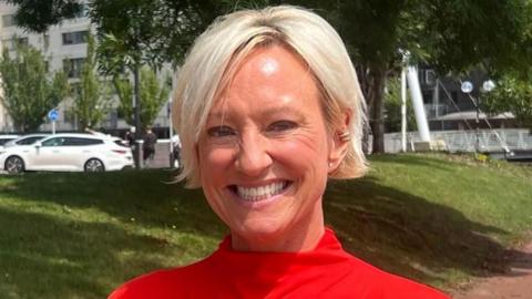 A woman wearing a red top with short blonde hair stands with buildings, a grassy verge and people visible in the background. She is smiling at the camera.