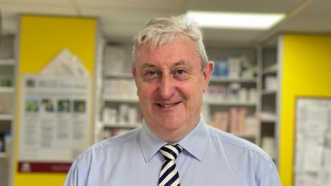 John Davey smiles at the camera, wearing a blue shirt and black and white striped tie, as he stands in a pharmacy 