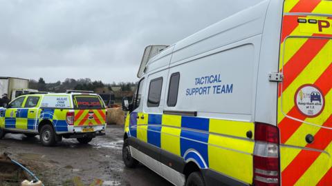 Two police tactical support vehicles are parked on what looks to be a farm. The ground is muddy with some puddles and there are fields and hay bales around them.