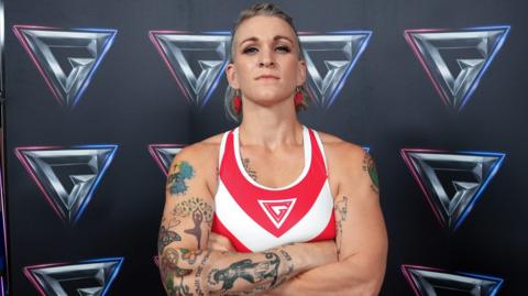 Rachael Phillips with short blonde hair, red earrings, a nose ring, a red and white sports top and tattoos on her arms.