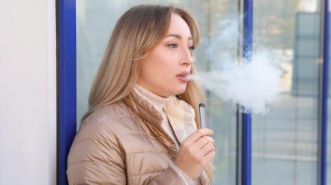 A woman with blue eyes and blonde hair blowing vapour from a grey e-cigarette held in her hand. She is wearing a light brown puffer jacket and a pink turtle neck top. The background includes a glass door and a white wall directly behind her.