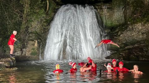 There is a large waterfall in the background with people in the pool in front of it wearing red hats and t-shirts