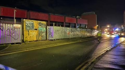A police cordon around a disused commercial building in Nottingham. The building, once a Staples office supplies shop, is boarded up with metal fencing. A number of fire service vehicles can be scene in the background while a firefighter crosses the road. It is night and dark outside. Blue lights can be scene from emergency service vehicles.