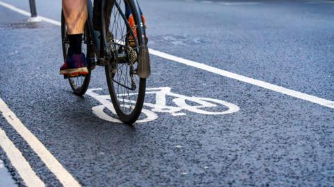 A cyclist riding in a designated bike lane marked by white lines and a painted bicycle symbol on a road. Only the bike’s rear wheel and the rider’s legs and shoes are visible.