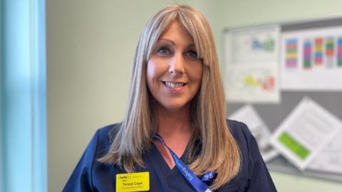 Teresa Cope has long blonde straight hair with a parted fringe. She is smiling and wearing dark blue scrubs with a blue lanyard around her neck and a yellow name badge that says "hello my name is Teresa Cope, chief executive officer". She is standing in an office with a pinboard on the right.