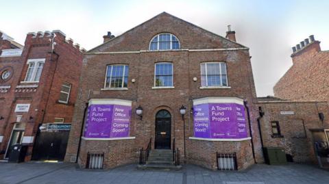 Four-storey brick building which slanted room. Bottom windows have purple signs up saying "A Towns Fund Project". 