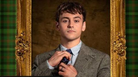 The image shows Tom Daley in a formal suit and tie, adjusting his tie. He is framed by an ornate golden frame with green tartan-patterned sides. The background features a textured brown surface.
